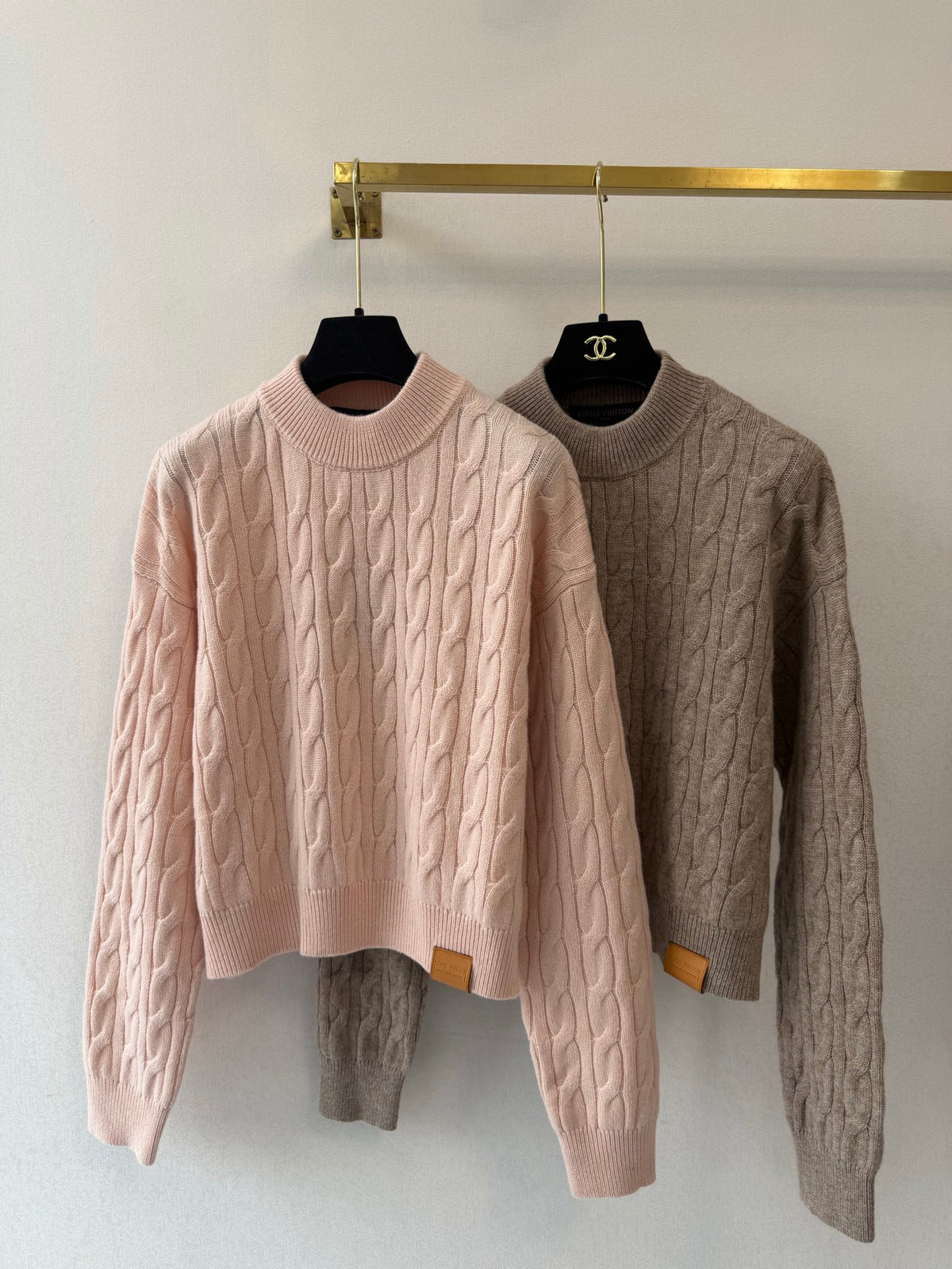 Two cable-knit crewneck sweaters hanging on black hangers from a gold clothing rack against a white wall. The sweater on the left is light pink, and the sweater on the right is taupe-brown.