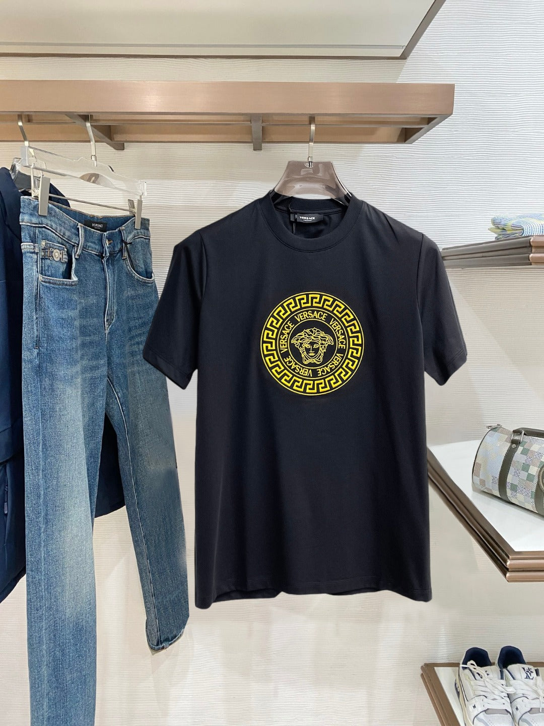 A black short-sleeve t-shirt with a large gold Versace Medusa logo on the chest, hanging on a wooden hanger in a retail store next to blue jeans.